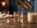 Steel pitcher pouring wax into a wicked glass jar between an empty prepared jar and a finished cream candle