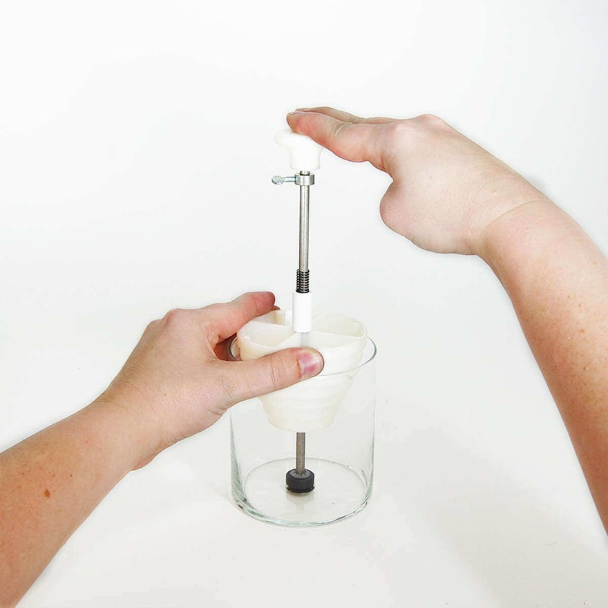 Hands pressing a wick-setting tool down into the bottom of a clear glass candle jar