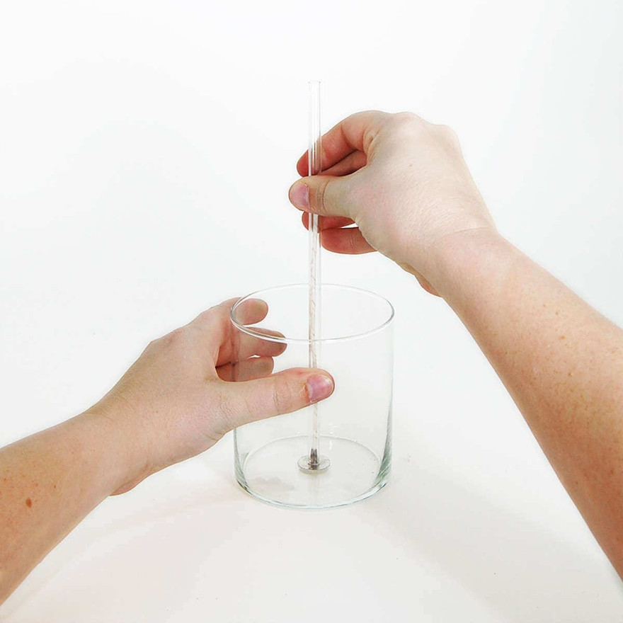 Hands lowering a wick-setting tool to center a wick tab in the bottom of a glass jar