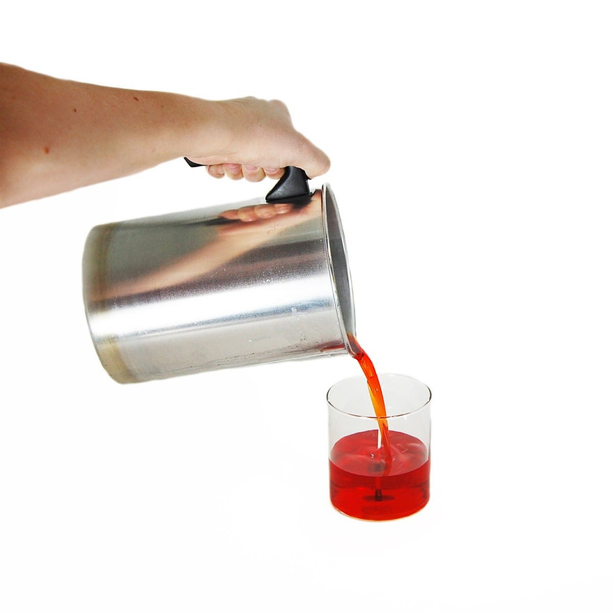 Hand pouring red-orange melted wax from a metal pitcher into a clear glass candle jar