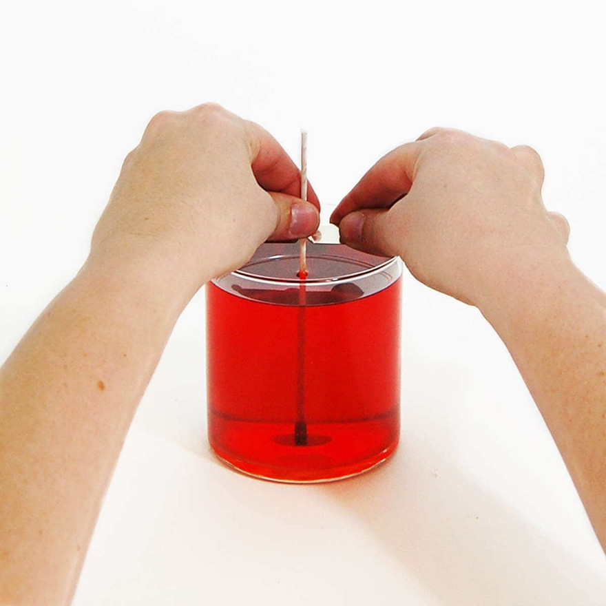 Hands steadying an upright wick in a clear glass jar filled with red liquid wax