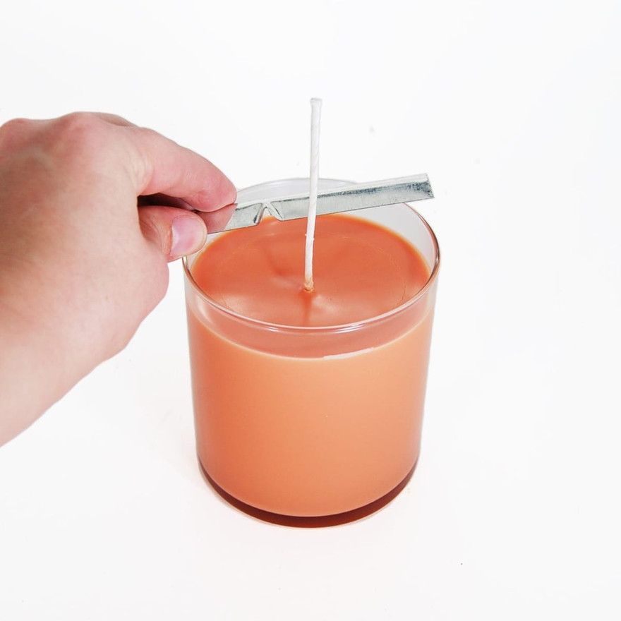 Hand resting a metal wick centering bar across a glass jar of setting orange candle wax