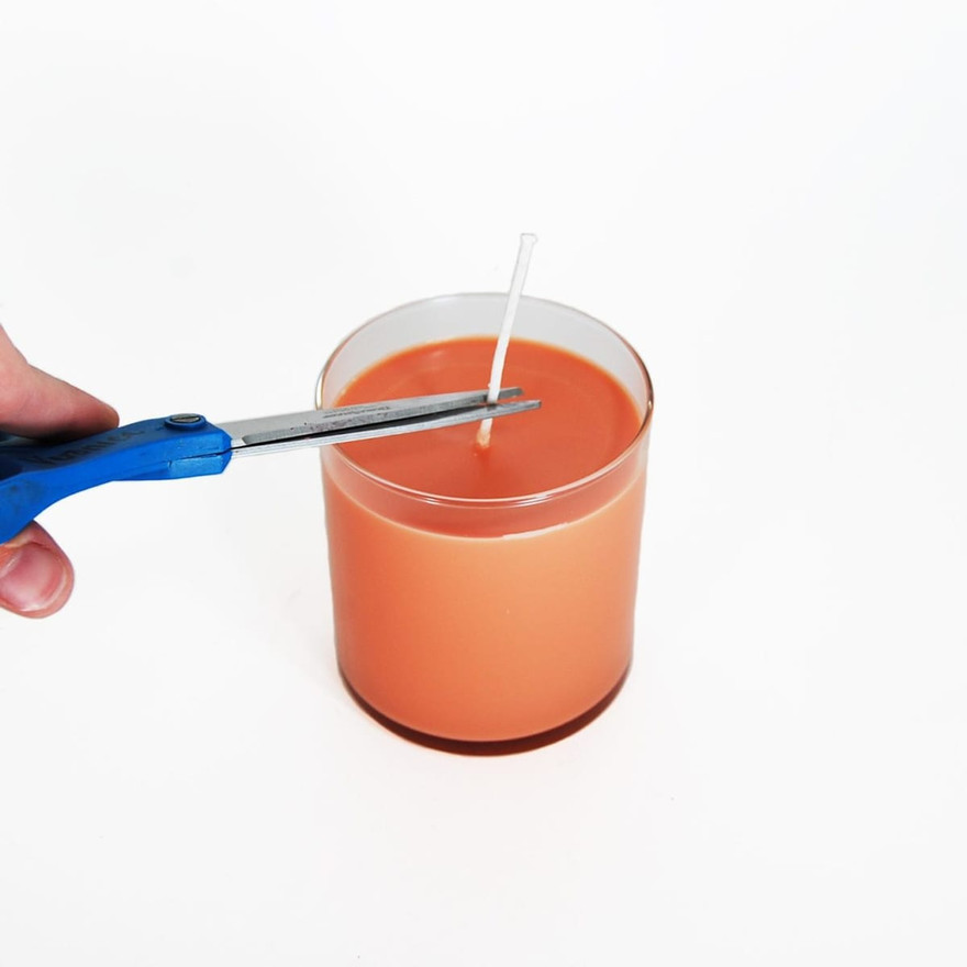 Blue-handled scissors trimming the wick on a finished orange candle in a glass jar