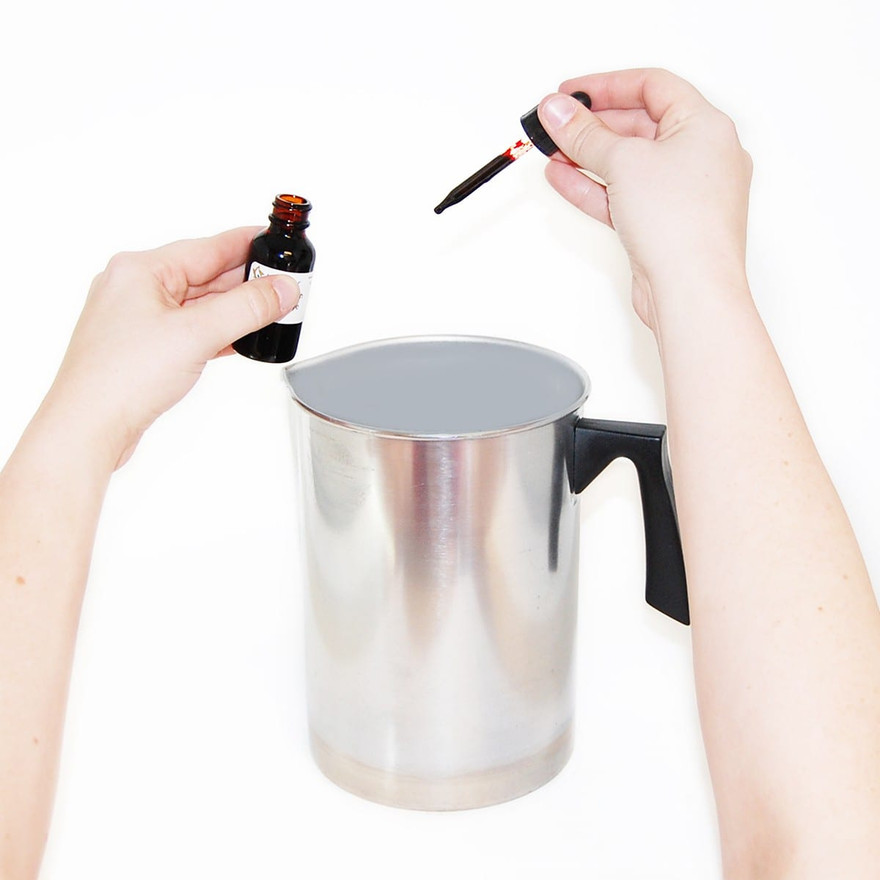 Hands holding a dropper of red dye and an amber bottle over a metal pitcher of melted wax