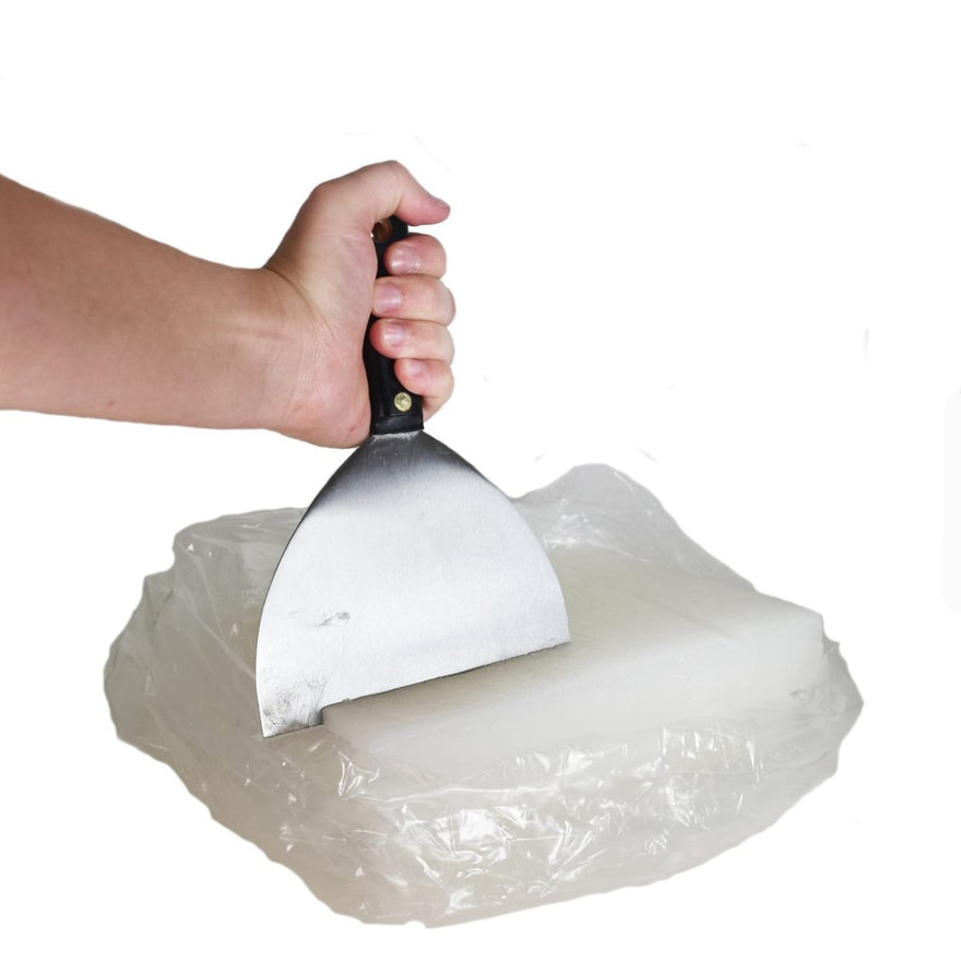 Hand pressing a steel putty knife into a plastic-wrapped slab of white wax