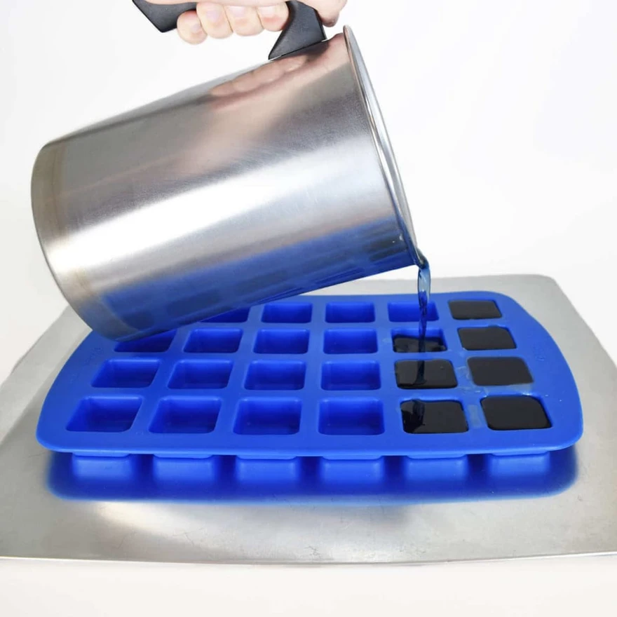 Dark blue wax pouring from a steel pitcher into the cavities of a blue silicone square mold