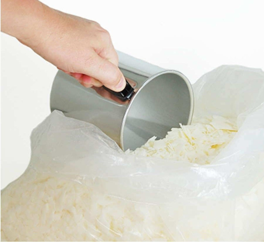 Hand scooping white soy wax flakes from an open bag with a metal pouring pitcher