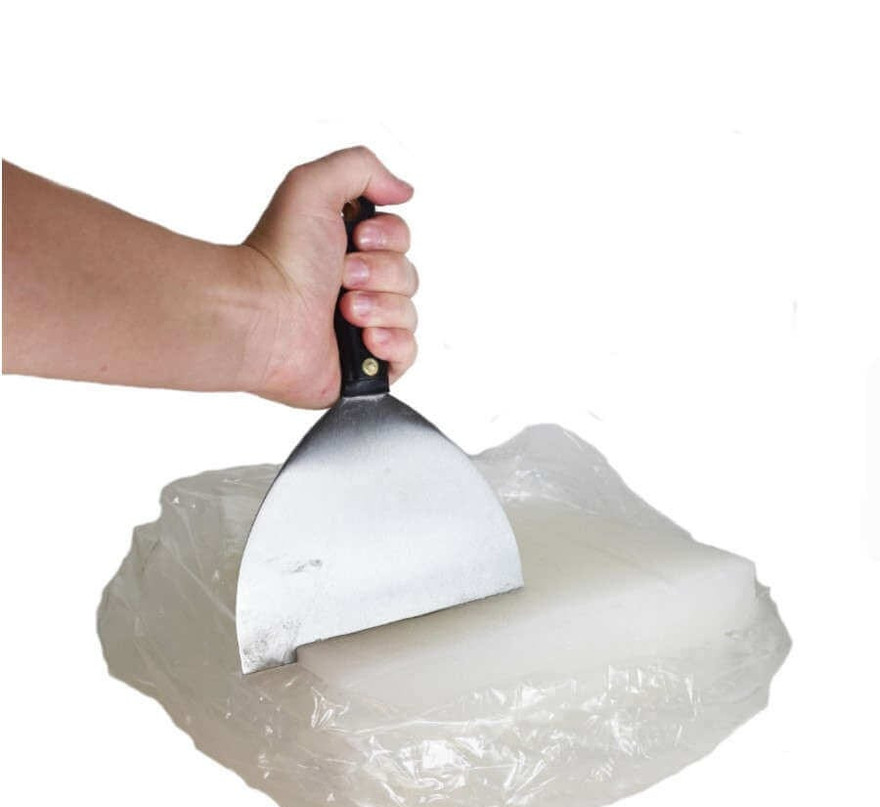 Hand pressing a wide putty knife into a bagged white wax slab to cut it