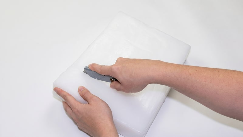 Two hands scoring a bagged white wax slab with a gray utility tool to snap a piece