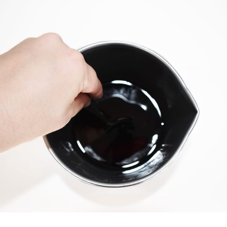 Overhead view of a hand stirring dark navy melted wax in a metal pouring pitcher